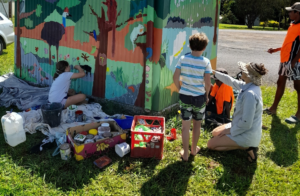 People painting a bus stop with images of animals and trees. The group of people are surrounded by paint cans.